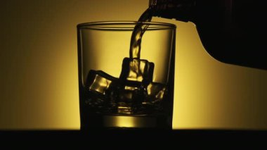 Sarı arka planda altın viski akıyor. Şişeden çıkan buzlarla bardağa alkol dökülüyor. Kahverengi şişeden akan viskinin yakın çekimi.