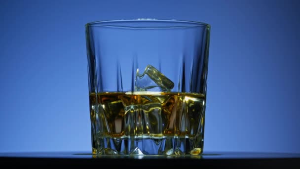 ウィスキーのバーボンと氷 青い背景にウイスキーの回転ガラス アルコール飲料 カクテル アルコール ガラス回転中の氷のキューブとバーボン Uhdのクローズアップ — ストック動画