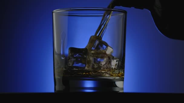 Whisky gegoten in glas op blauwe achtergrond. Glas whisky met ijsblokjes — Stockvideo