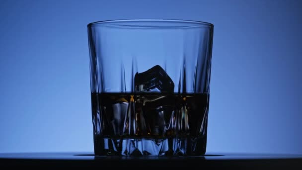 Whiskey bourbon with ice. Rotated glass of whisky on blue background in 4K, UHD — 비디오
