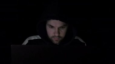 Kapüşonlu bilgisayar korsanı karanlık odada dizüstü bilgisayarla çalışıyor. Adam stresli ve etrafına bakıyor.