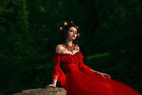 The lady in lush red dress with open shoulders. Around her neck is unusually luxurious decoration.The head decorated with a crown handmade.The fairy Princess was sitting on a rock, dreamy head thrown back.Fantastic shot. Fashionable toning. Creative