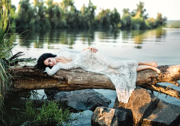 Роскошная брюнетка в белом платье изящно лежит на бревне — стоковое фото