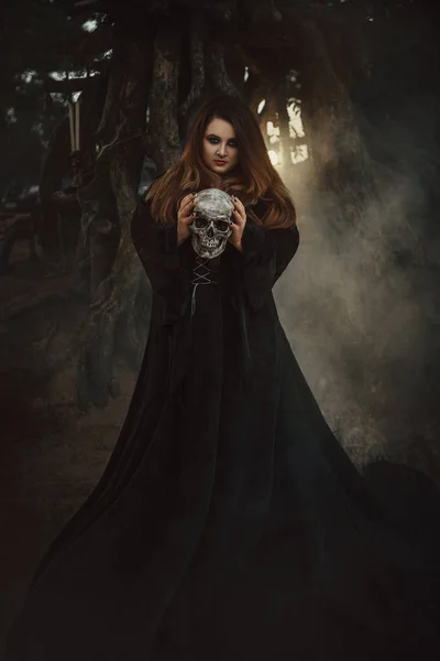 Une jeune femme en robe noire avec de longs cheveux regardant directement la caméra debout et tenant un crâne, un brouillard sombre, une illustration fabuleuse . — Photo