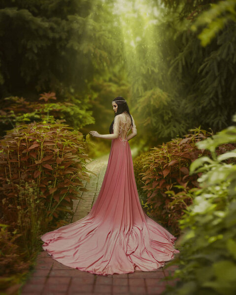The elf walks in the summer garden. A girl with long ears in a beautiful pink dress with an open back and with a long train. Artistic processing