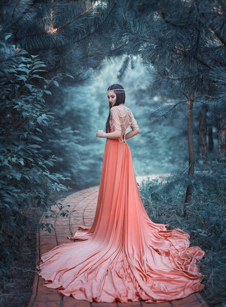 The elf walks in the summer garden. A girl with long ears in a beautiful orange dress with an open back and with a long train. Artistic processing