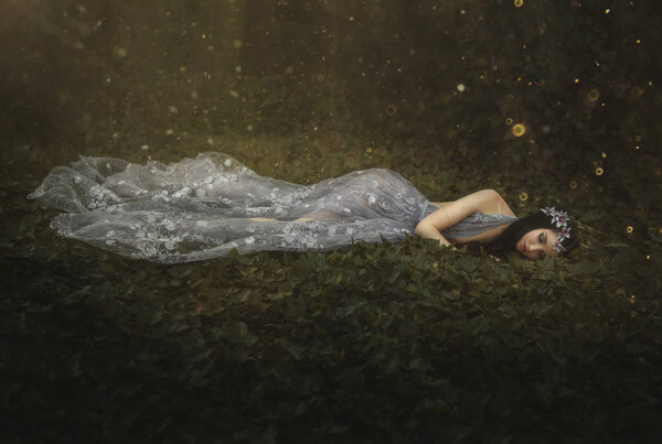 Sleeping Beauty. The girl lies on the grass in a dark, dense forest. An unusual transparent dress. Artistic processing