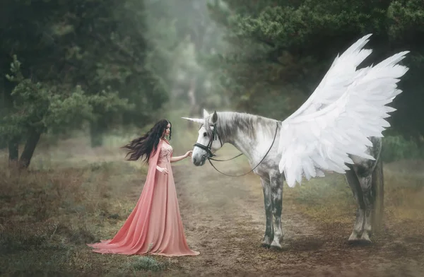 Hermosa, joven elfo, caminando con un unicornio en el bosque Ella está vestida con un vestido largo de color naranja con una capa. La pluma ondea maravillosamente en el viento. Fotografía artística —  Fotos de Stock