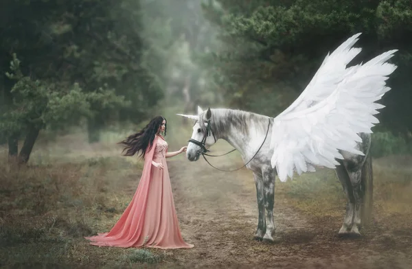 Hermosa, joven elfo, caminando con un unicornio en el bosque Ella está vestida con un vestido largo de color naranja con una capa. La pluma ondea maravillosamente en el viento. Fotografía artística Imágenes De Stock Sin Royalties Gratis