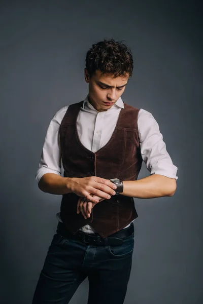 A young, handsome guy posing on a gray studio background. The man is stylish and elegantly dressed in jeans, a shirt and a brown vest. The guy is looking at his watch