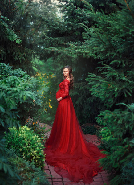 An elegant, pregnant woman walks in a beautiful garden in a luxurious, expensive red dress with a long train. Artistic Photo