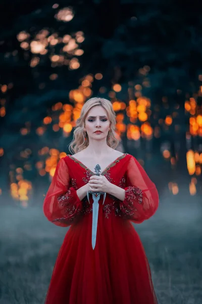 Beautiful, dangerous blonde queen in a red fashion lush dress with a dagger in her hands. Backdrop dark fantasy forest in a fog. Fiery sunset. Concept Revenge, conspiracy and betrayal. Halloween party — Stock Photo, Image