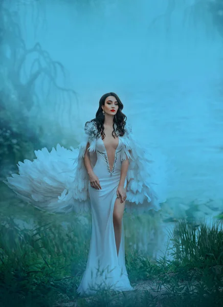 Strong attractive brunette woman in luxurious fluttering waving outfit. long Cloak with feathers and a white sexy dress deep neckline. Spring Haze over water by river. Autumn foggy weather. Green lake — Stock Photo, Image