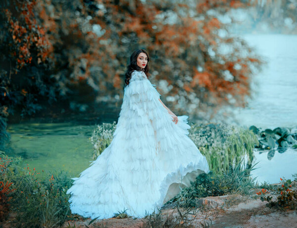 attractive brunette woman in luxurious fluttering waving outfit. A gentle snow queen walks. green lake. long Cloak with feathers. Spring Haze over water by river. Autumn foggy weather. Yellow trees
