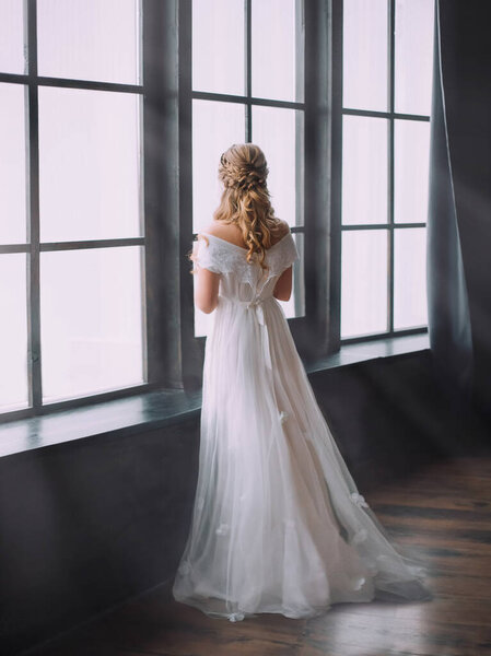 An attractive pensive blonde woman with a creative hairstyle for long hair is standing in a black gothic dark old room. Art photography. White wedding dress of the bride. Turned away, without face