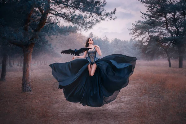 Attractive seductive sexy fantasy witch levitating in air. Woman with black raven on hand. wings bird. silk dress flutters. Gothic vintage style design. Cold winter forest nature backdrop with fog — Stock Photo, Image