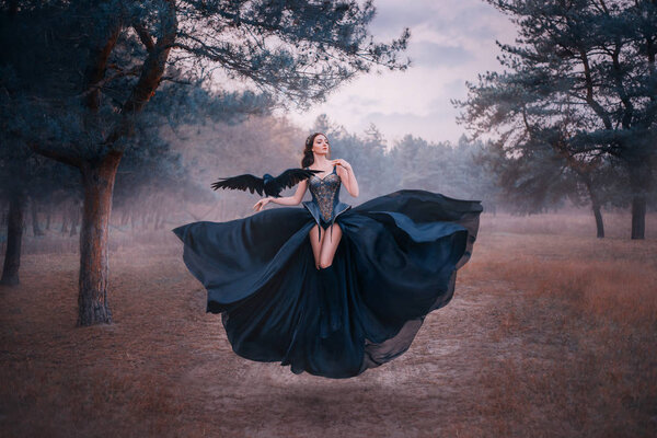 привлекательная сексуальная фантазийная ведьма левитирует в воздухе. Женщина с черным вороном на руке. крылья птицы. шелковое платье трепещет. Готический винтажный дизайн. Холодные зимние леса природа фон с туманом
