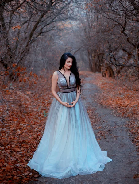 Donna bruna con lunghi capelli cammina nella foresta autunnale di novembre. Sfondo nero alberi nudi e foglie d'arancio cadute. La regina ama la natura. Reale lusso gonfio abito turchese. Immagine del partito di laurea — Foto Stock