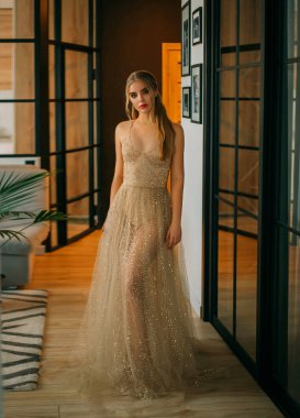 Şık, alımlı, pırıl pırıl, altın rengi şeffaf elbiseli sarışın kadın. Moda tarzı. Parti için lüks bir imaj. Mezuniyet için akşam kıyafeti, Noel, yeni yıl. Arkaplan iç tavan arası