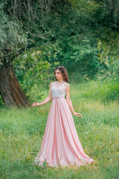 Cute attractive brunette woman enjoying nature in delicate elegant pink silk dress with white lace top. Image for party graduation prom ball stylish evening outfit celebration. Fashion glamor summer