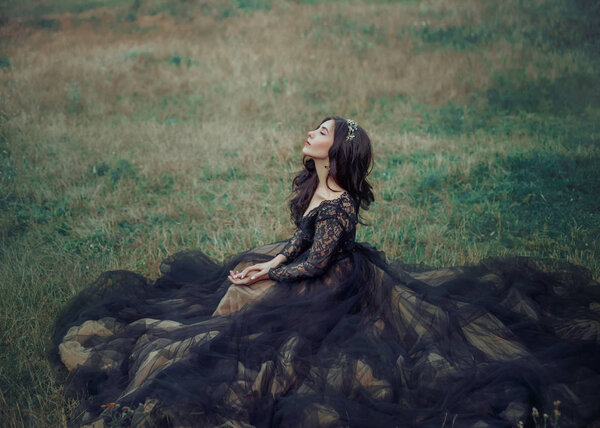 Cute lady in black elegant puffy dress sits on green grass. eyes closed, enjoys nature. Brunette woman with long wrap hair. stylish volumetric hairstyle. Gothic metal crown hoop fashion couture design