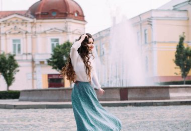 Esmer kadın öğrenci dans ediyor, sokak şehrinin etrafında dönüyor. Uzun dalgalı saçlar hareket halinde. Neşeli mutlu kız. Gülümse. Beyaz eski bir bluz, naneli bir etek. Çiçek izi.