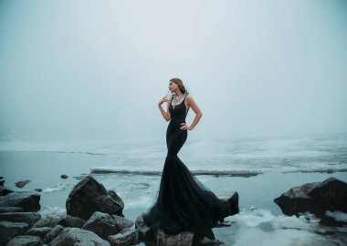 Seksi uzun siyah elbiseli siluet denizkızı kuyruğu balığı. Arka planda nehir buzlu su soğuk kar taşları. Kış donmuş göl. Karanlık Gotik Kraliçe. Moda tasarımı yaratıcı kolye tasması boncukları.