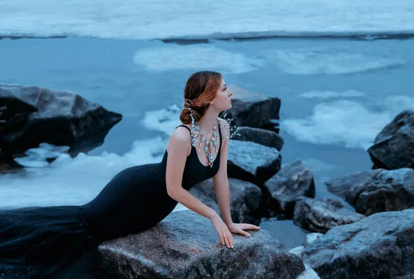Vörös hajú hableány lány ül a parton fagyott folyó várja a visszajárót. a szerelem fogalmát. Fekete luxus elegáns testalkatú ruha. Kreatív ékszerek nyaklánc drágakövek gyémánt ezüst gyöngyök — Stock Fotó