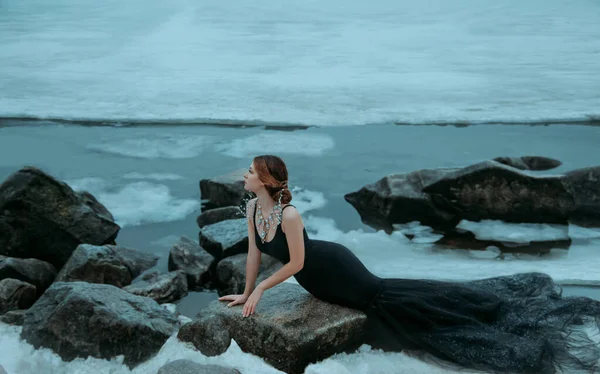 Mujer sexy vestido negro largo silueta sirena sentada en piedra. telón de fondo río hielo agua fría. Lady Elegante reina del mar disfrutando del invierno. Joyería creativa collar de piedras preciosas cuentas de collar de lujo real —  Fotos de Stock