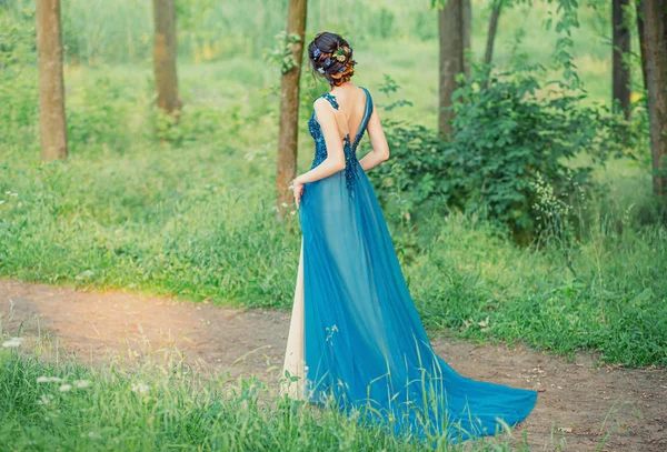 Einsame Mädchen gehen grünen Baum Wald blaues Abendkleid. Haar verziert blaue Blumen Kornblumen. elegante brünette Frisur. Blick nach hinten geöffnet. sonniger heller Tag Sommer Frühling. Abschlussball Party Ballkunst — Stockfoto