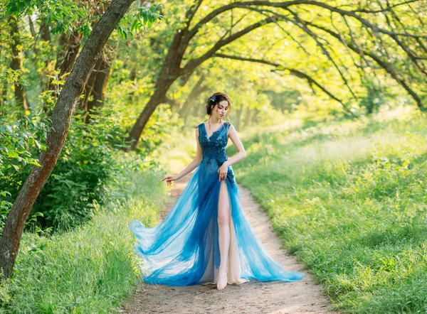 Eenzaam sexy meisje naakt been lopen gebladerte groen bos avond chique jurk. haar versierde blauwe bloemen van korenbloemen. Elegant brunette kapsel. Zonnige heldere dag. Zomer lente. bruiloftsfeest. van kunst — Stockfoto
