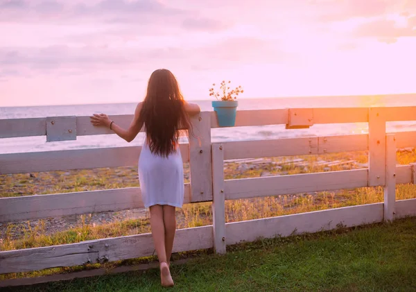 Vista posterior morena Chica de lujo pelo largo, vestido sexy transparente. Fondo soleado amarillo brillante puesta del sol valla de madera. Vacaciones en el mar. turista disfrutar del sol brillante verano. arte fantasía naturaleza Georgia — Foto de Stock