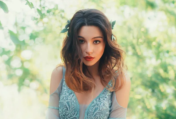 Gros plan portrait jolie jeune fille brune avec les cheveux bouclés ondulés lâches. Coiffure décorée feuilles vertes. Nymphe en dentelle robe aquarelle bleue. Soirée Maquillage cosmétiques naturels. toile de fond art soleil éblouissement — Photo