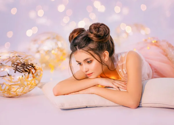 Dremy girl with red hair an elegant hairstyle two buns evening makeup lies on pillow hugging bright glowing ball. Model in delicate pink dress. Background fantastic sparkles golden balloons birthday — Stock Photo, Image