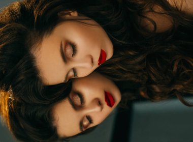Model aynanın gözleri kapalı yatar. Yakın plan portre prensesi. Parlak gece, kırmızı dudaklar kahverengi parlak gölgeler oluşturur. Zarif dalgalı saç stili, uzun, sağlıklı saçlı esmer. yüz yansıtma arkaplanı