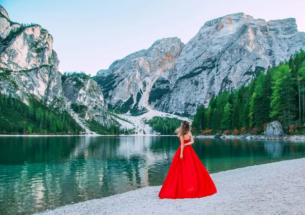 Princesa pelo rubio volar en el viento. vestido largo de seda roja vintage rechazado se encuentra en la orilla esmeralda fabuloso lago Braies. Chica disfruta de la belleza misteriosa naturaleza dolomita blanco montañas alpino verde bosque — Foto de Stock