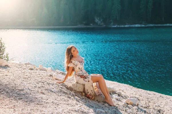 Encantadora mujer se sienta orilla lago alpino disfruta del sol fantástica naturaleza verde bosque silencio relajarse. Chica con el pelo largo que fluye vestido blanco de seda piernas desnudas sexy. Árboles de telón de fondo agua ondulada turquesa — Foto de Stock