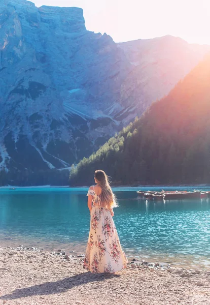 Chica con el pelo volando en el viento de pelo rojo, vestido de seda larga elegancia estampado floral dio la vuelta se levanta en la orilla del lago Bryce disfrutar del silencio belleza sol montañas. Fondo barco agua montaña con bosque — Foto de Stock