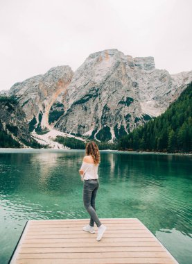 Kızıl saçlı, uzun saçlı, arkasını dönmüş, Braies Gölü 'nün ahşap iskelesinde duruyor. Kadın çıplak, omuz üstü, beyaz, havalı, seksi bluz mavi pantolon. Arkaplan muhteşem manzara dağlar yeşil orman temiz su