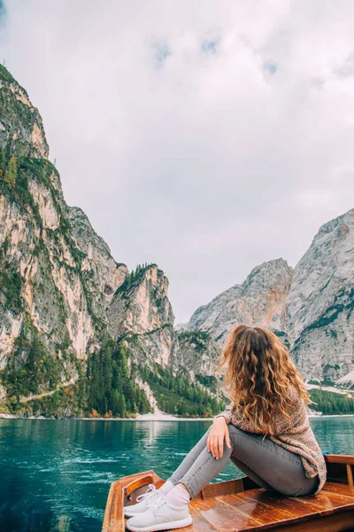 Mujer turista joven, pelo largo y ondulado se volvió atrás se sienta en viaje en barco de madera. Ropa casual cálido suéter marrón disfrutar del silencio sueño relajación lujo naturaleza italiano Alpes telón de fondo fabuloso lago Braies — Foto de Stock