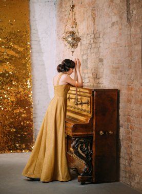 Beautiful Great Gatsby woman holds hands on piano prays no face with finger wave hairstyle in long luxurious gold dress retro style 30. Backdrop brick wall bright sparkle screen. Party Prom Christmas