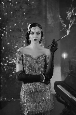 Closeup vertical image portrait young woman with mouthpiece in hand cigarette smoke on backdrop brilliant screen piano burning candle in black and white color. Finger wave hairstyle fashion style 1920