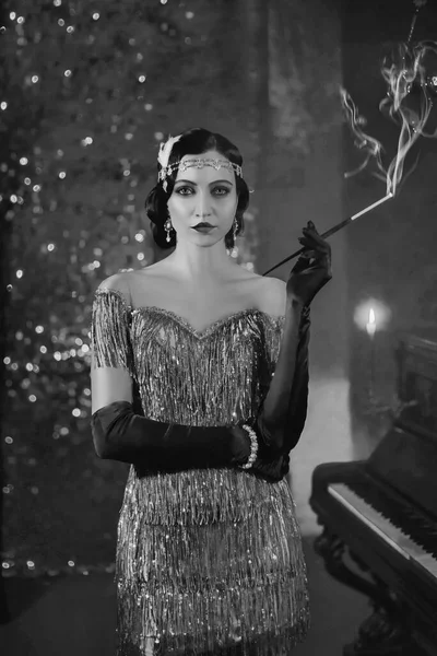 Closeup vertical image portrait young woman with mouthpiece in hand cigarette smoke on backdrop brilliant screen piano burning candle in black and white color. Finger wave hairstyle fashion style 1920 — Stockfoto