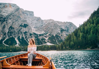 Beautiful redhaired girl clothes in white silk sexy blouse long sleeves bare shoulders gray trousers sitting in boat. Tourist enjoy view alpine lake of Braies. Backdrop Italian mountains green forest