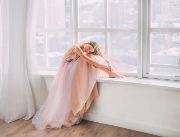 Umění spící krásu snů. Blond mladá žena s dlouhými splývajícími vlasy. Sedí na okenních parapetech, objímá nohy. Vinobraní pokoj bílé okno. Princezna roztomilá tvář. luxusní pastelová béžová fialová maturitní šaty — Stock fotografie