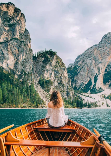 Redhaired girl turned away, sitting in the boat. Tourist in white blouse bare shoulders enjoy beauty alpine lake Braies backdrop magical nature dolomite mountains covered green forest. Vertical image