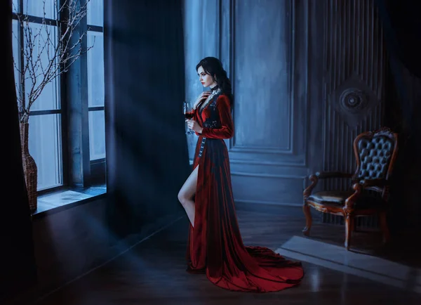 Linda jovem mulher sexy vampiro no castelo escuro medieval. Vestido gótico longo vermelho. Cabelo ondulado preto. Fundo vintage quarto interior. Segure vinho de sangue de vidro. imagem rainha do feriado de terror da noite halloween — Fotografia de Stock