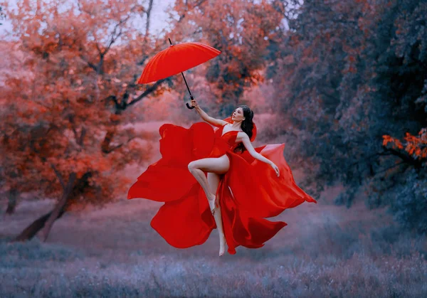 Όμορφη κομψή ευτυχισμένη γυναίκα πετούν με ομπρέλα. Κόκκινο μακρύ σέξι μεταξωτό φόρεμα ύφασμα φτερουγίζει στον άνεμο. Θεά που χορεύει χαρούμενη στον αέρα. Μυστικιστικά φθινοπωρινά δέντρα. Τέχνη φαντασία αιώρηση πριγκίπισσα — Φωτογραφία Αρχείου
