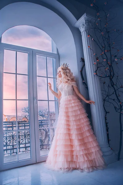 Fantasie mooie middeleeuwse vrouw prinses staat in de buurt van het raam kasteel witte kamer. Meisje op zoek naar buiten met hoop. Roze lange luxe jurk. Lady Queen golvend blond haar. Vintage koninklijke kroon. Blijf thuis. — Stockfoto