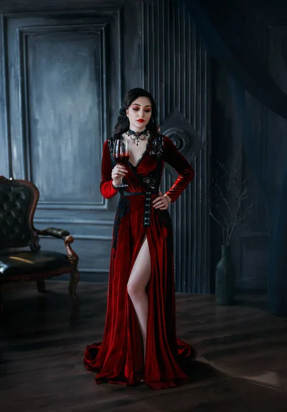 Ein mittelalterlicher junger Vampir hält ein Glas blutigen Weins in der Hand. Rote lange sexy Kleid Schleppe. Gothic dark quuen Dame. Schwarze lange wellige Haare sexy nacktes Bein. Hintergrund Vintage Night Room Interieur. halloween — Stockfoto